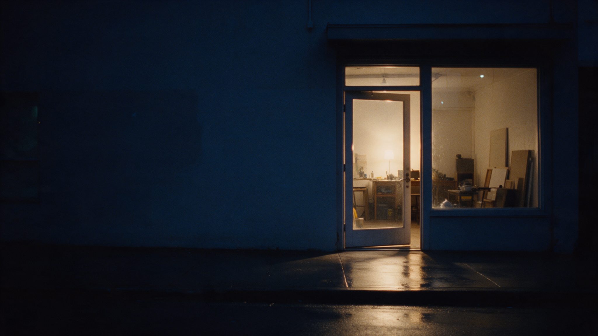 The open door of a small business at night, warm light on the wet sidewalk.